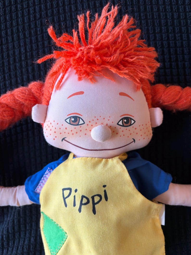 May include: A soft doll with bright red yarn hair in braids and a top knot. The doll has a smiling face, freckles, and is wearing a yellow top with the name "Pippi" printed on it. The doll's shirt has blue sleeves.