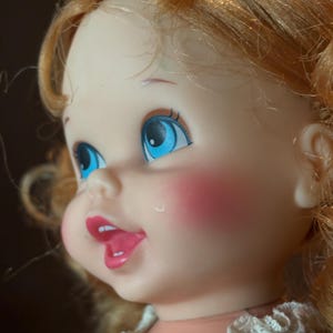 May include: A vintage doll with blue eyes, rosy cheeks, and open mouth. The doll has blonde, curly hair and is wearing a dress with a white lace collar. The doll's face is turned to the side, showcasing its features.