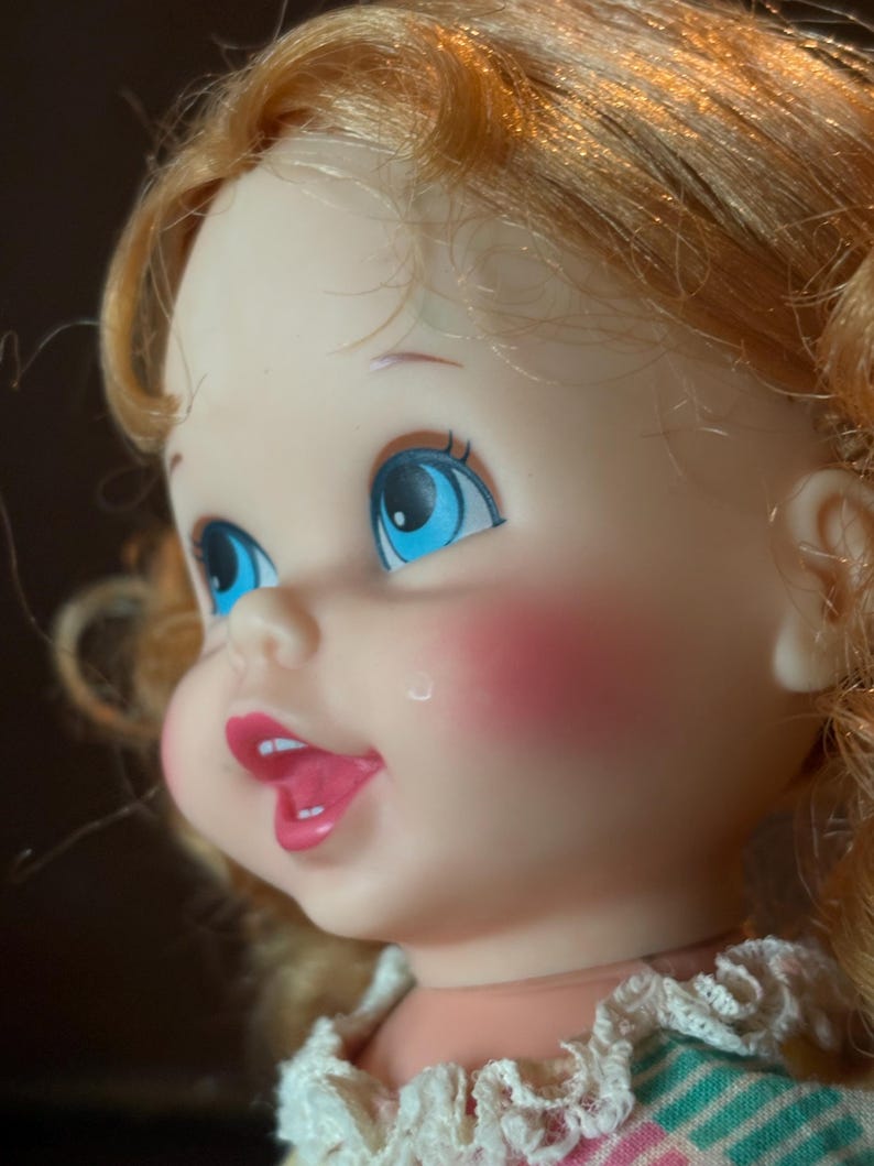 May include: A vintage doll with blue eyes, rosy cheeks, and open mouth. The doll has blonde, curly hair and is wearing a dress with a white lace collar. The doll's face is turned to the side, showcasing its features.