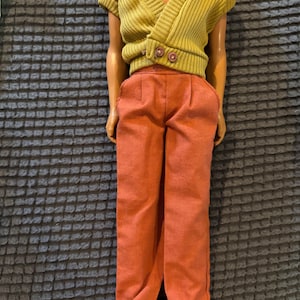 May include: A doll dressed in a mustard yellow wrap-style sweater with two brown buttons and rust-coloured trousers. The trousers have a pleated front and a wide-leg design. The doll is on a dark grey textured surface.