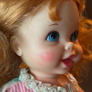 May include: A vintage doll with red hair and blue eyes. The doll has rosy cheeks and is wearing a dress with a white lace collar. The dress has pink, yellow, and blue stripes. The doll's mouth is open in a slight smile.
