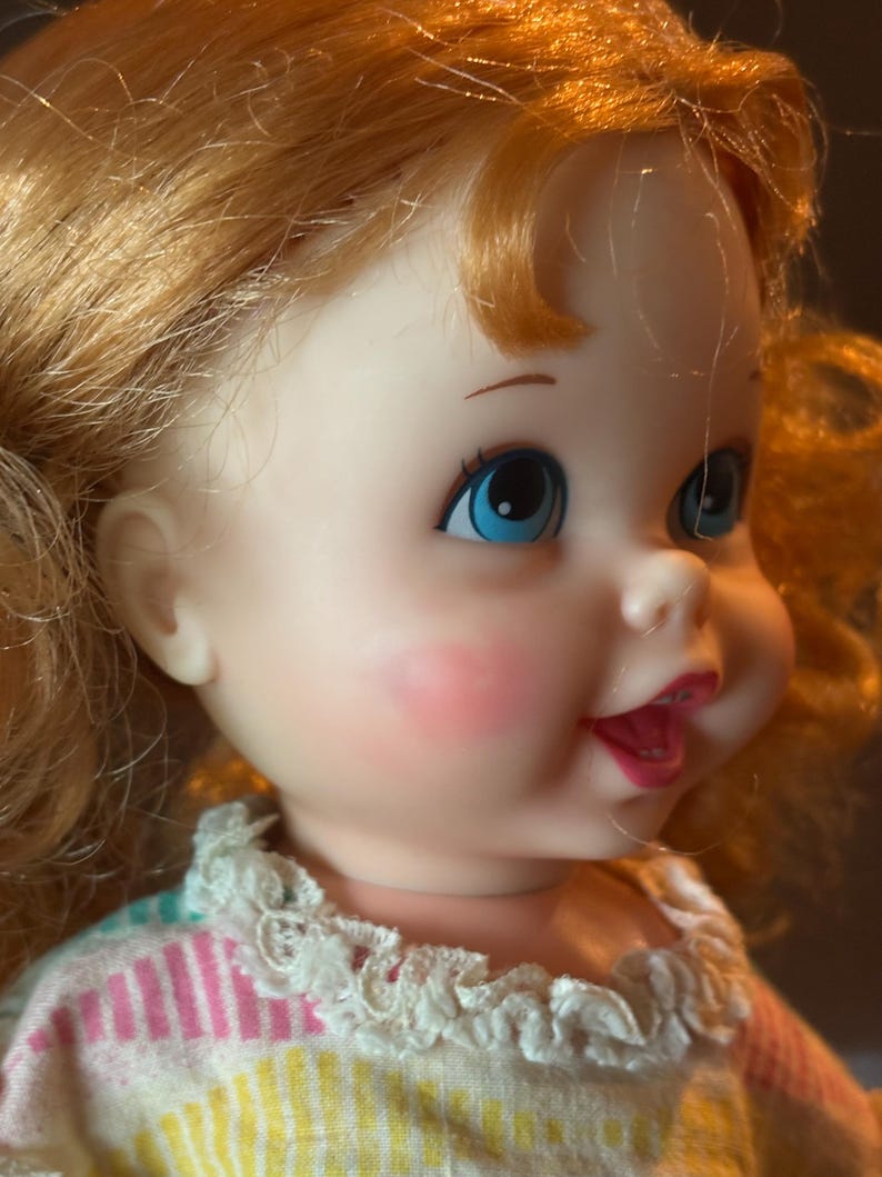 May include: A vintage doll with red hair and blue eyes. The doll has rosy cheeks and is wearing a dress with a white lace collar. The dress has pink, yellow, and blue stripes. The doll's mouth is open in a slight smile.