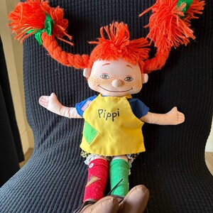 May include: A handmade doll with bright red yarn pigtails, each tied with a green ribbon. The doll wears a yellow top with the name "Pippi", blue sleeves, and mismatched socks. Brown shoes complete the outfit.