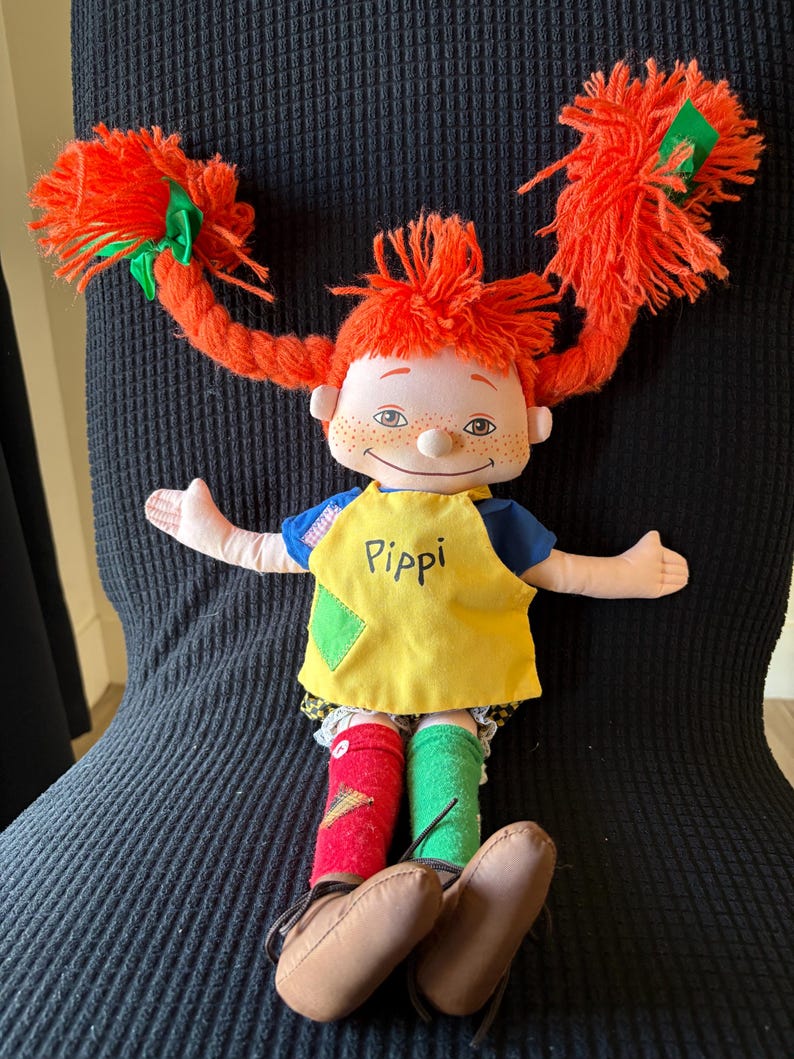 May include: A handmade doll with bright red yarn pigtails, each tied with a green ribbon. The doll wears a yellow top with the name "Pippi", blue sleeves, and mismatched socks. Brown shoes complete the outfit.