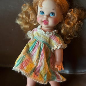 May include: A vintage doll with blonde curly pigtails and blue eyes. The doll wears a colorful striped dress with a white lace trim. The doll has rosy cheeks and a small open mouth.