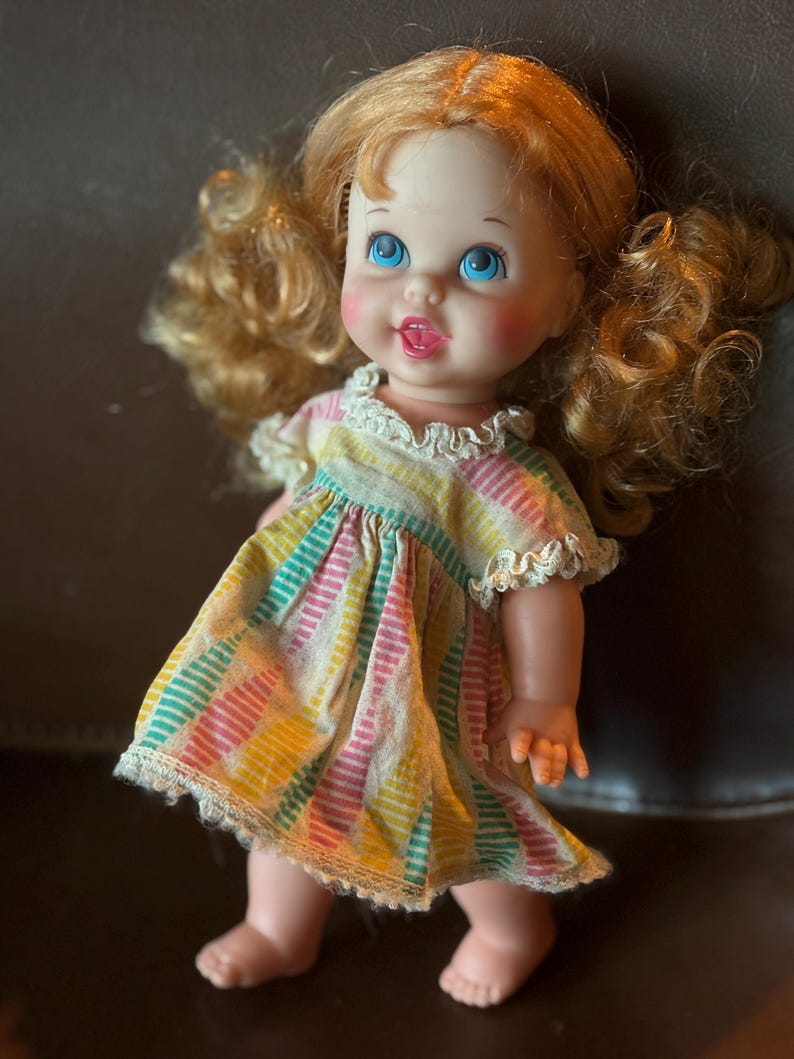 May include: A vintage doll with blonde curly pigtails and blue eyes. The doll wears a colorful striped dress with a white lace trim. The doll has rosy cheeks and a small open mouth.