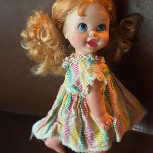 May include: A vintage doll with red curly hair styled in pigtails. The doll has blue eyes, rosy cheeks, and a smiling expression. It wears a colorful striped dress with lace trim. The doll is standing on a brown surface.