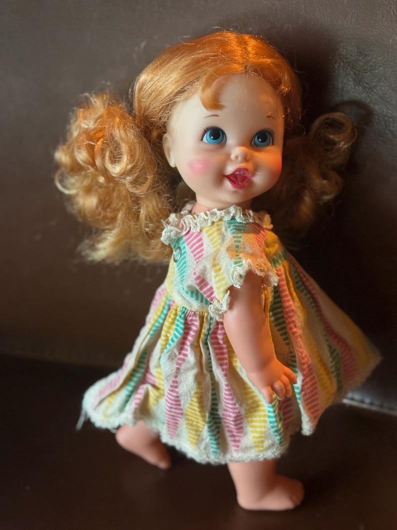 May include: A vintage doll with red curly hair styled in pigtails. The doll has blue eyes, rosy cheeks, and a smiling expression. It wears a colorful striped dress with lace trim. The doll is standing on a brown surface.