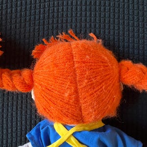 May include: A doll with vibrant orange yarn pigtails and a blue dress with yellow straps. The doll's hair is styled in two thick braids, and the dress has a white collar. The doll is a children's toy.