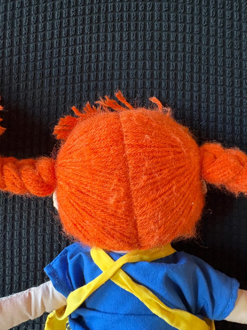 May include: A doll with vibrant orange yarn pigtails and a blue dress with yellow straps. The doll's hair is styled in two thick braids, and the dress has a white collar. The doll is a children's toy.
