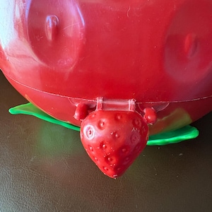 Pode incluir: Um recipiente vermelho de plástico em forma de morango com uma base de folha verde. O recipiente tem um pequeno fecho em forma de morango. O recipiente está sobre uma superfície castanha.