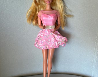 May include: A vintage doll with long blonde hair, blue eyes, and a pink headband. The doll wears a pink dress with a sheer neckline and a floral-patterned skirt. Pink shoes complete the outfit. The doll is a classic toy.