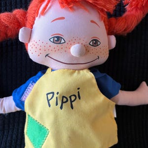 May include: A soft doll with bright red pigtails, a smiling face, and freckles. The doll is dressed in a blue top and a yellow apron with the name "Pippi" printed on it. A green diamond patch is sewn on the apron.