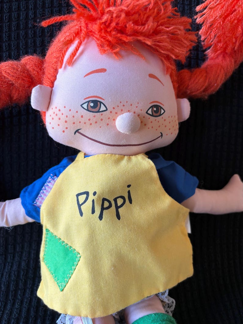 May include: A soft doll with bright red pigtails, a smiling face, and freckles. The doll is dressed in a blue top and a yellow apron with the name "Pippi" printed on it. A green diamond patch is sewn on the apron.