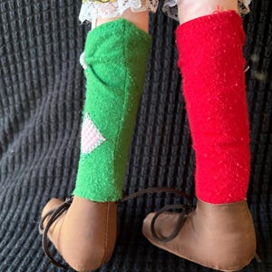 May include: Close-up of a doll's legs. The doll wears brown shoes with laces, green and red socks, and yellow and black checkered trousers with white lace trim. A pink and white patch is sewn on the green sock.