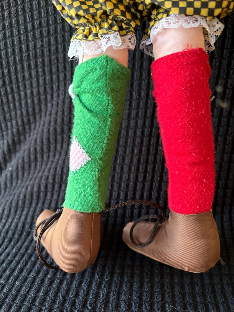 May include: Close-up of a doll's legs. The doll wears brown shoes with laces, green and red socks, and yellow and black checkered trousers with white lace trim. A pink and white patch is sewn on the green sock.