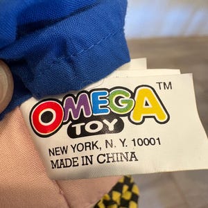 May include: A close-up of a toy tag with the brand name "OMEGA TOY" in colourful letters. The tag also includes the text "NEW YORK, N. Y. 10001" and "MADE IN CHINA". A blue fabric is visible at the top.