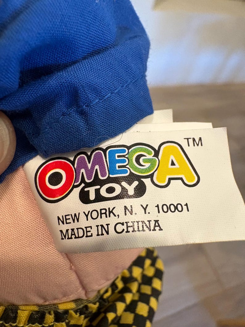 May include: A close-up of a toy tag with the brand name "OMEGA TOY" in colourful letters. The tag also includes the text "NEW YORK, N. Y. 10001" and "MADE IN CHINA". A blue fabric is visible at the top.