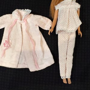 May include: A doll wearing a white pajama set with small pink dots and lace trim, accompanied by a separate light pink robe featuring a white lace collar, long sleeves, two pink ribbon ties, and a small embroidered pink cat on the pocket.