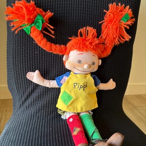 May include: A Pippi Longstocking doll with vibrant orange pigtails, each secured with a green bow. The doll is dressed in a yellow apron with "Pippi" written on it, a blue top, and mismatched red and green socks. Brown shoes complete the outfit.