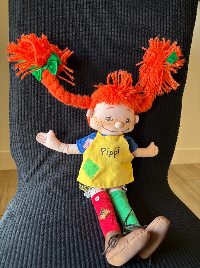 May include: A Pippi Longstocking doll with vibrant orange pigtails, each secured with a green bow. The doll is dressed in a yellow apron with "Pippi" written on it, a blue top, and mismatched red and green socks. Brown shoes complete the outfit.