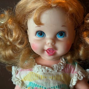 May include: A vintage doll with bright blue eyes, rosy cheeks, and a smiling expression. The doll has curly, light-red hair and wears a dress with a colorful striped pattern and ruffled trim. A classic toy.