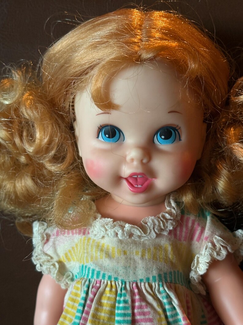 May include: A vintage doll with bright blue eyes, rosy cheeks, and a smiling expression. The doll has curly, light-red hair and wears a dress with a colorful striped pattern and ruffled trim. A classic toy.