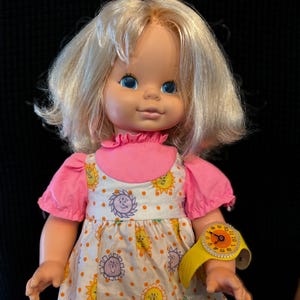 Puede incluir: Una muñeca vintage rubia con un vestido rosa y blanco con un estampado de sol amarillo. La muñeca tiene ojos azules y lleva un reloj de plástico amarillo con una esfera blanca.