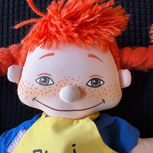 May include: A soft doll with bright orange yarn hair in pigtails. The doll has a white nose, freckles, and brown eyes. It wears a yellow bib with the name "Pippi" printed in black. The doll's shirt is blue.