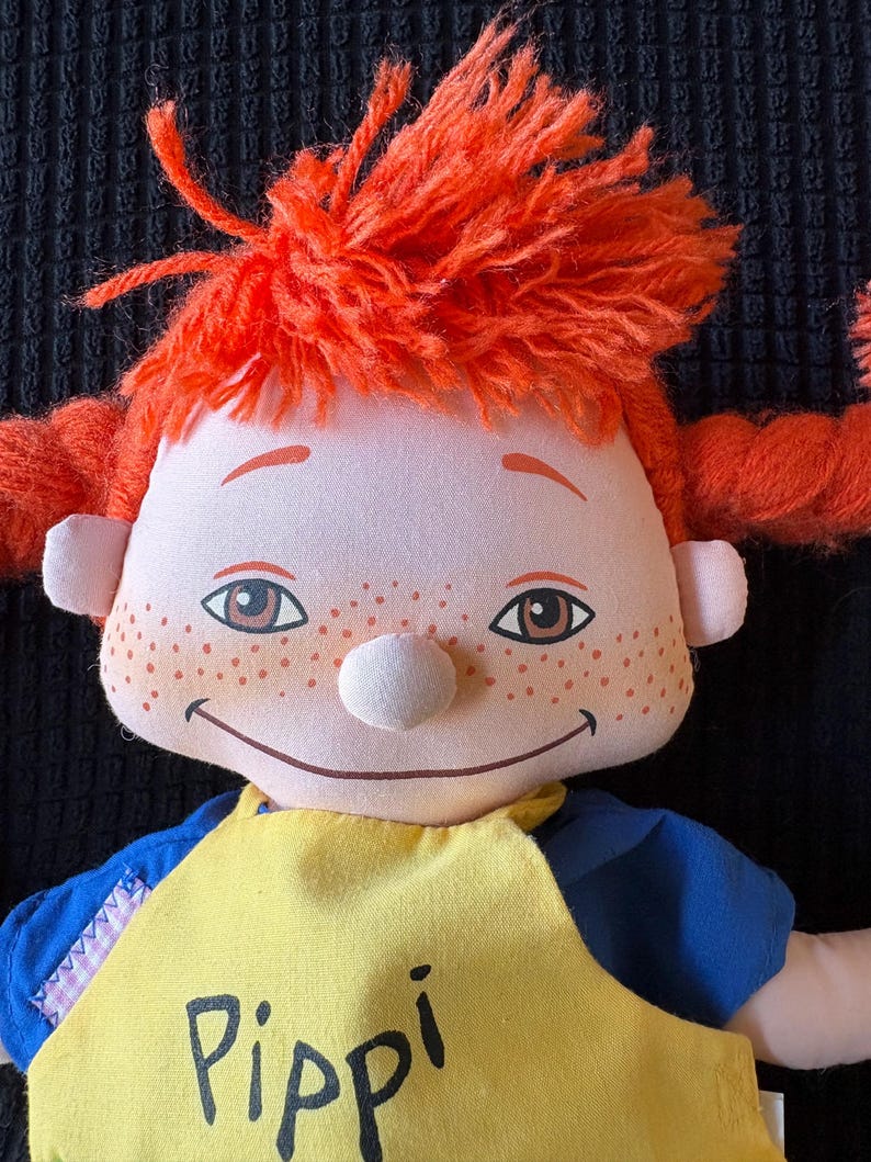 May include: A soft doll with bright orange yarn hair in pigtails. The doll has a white nose, freckles, and brown eyes. It wears a yellow bib with the name "Pippi" printed in black. The doll's shirt is blue.