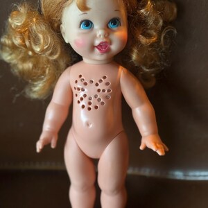 May include: A vintage doll with light skin, blue eyes, and curly blonde pigtails. The doll has rosy cheeks and a small, open mouth with a pink tongue. The doll's chest features a heart-shaped pattern of holes.