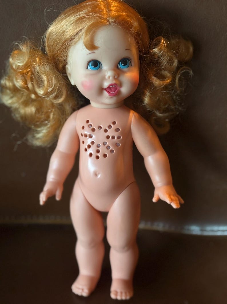 May include: A vintage doll with light skin, blue eyes, and curly blonde pigtails. The doll has rosy cheeks and a small, open mouth with a pink tongue. The doll's chest features a heart-shaped pattern of holes.