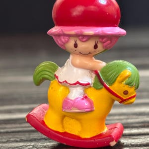 May include: A red and pink Strawberry Shortcake doll riding a yellow rocking horse with green leaves. The doll is wearing a white dress with pink shoes.