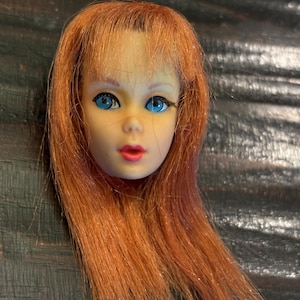 Op de afbeelding: Een vintage poppenhoofd met lang, roodbruin haar en helderblauwe ogen. Het gezicht van de pop heeft geschilderde roze lippen en delicate wimpers. Het hoofd is gemaakt van een licht materiaal, waarschijnlijk plastic, en is een verzamelobject.