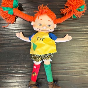May include: A Pippi Longstocking doll with vibrant orange yarn pigtails, each adorned with a green bow. The doll is dressed in a yellow top with "Pippi" printed on it, black and white shorts, and mismatched red and green socks. Brown shoes complete the outfit.