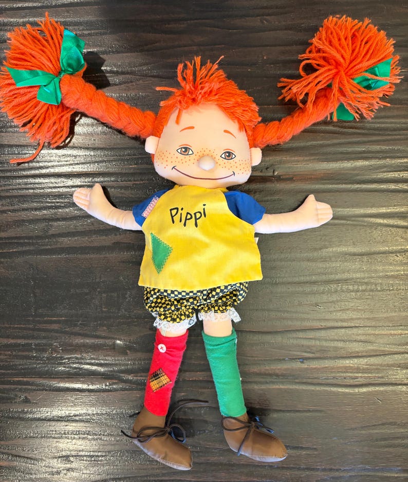 May include: A Pippi Longstocking doll with vibrant orange yarn pigtails, each adorned with a green bow. The doll is dressed in a yellow top with "Pippi" printed on it, black and white shorts, and mismatched red and green socks. Brown shoes complete the outfit.