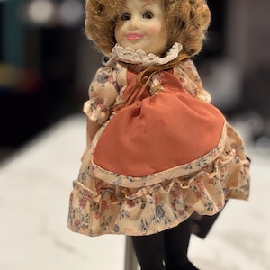 Könnte beinhalten: Vintage Puppe mit bemaltem Gesicht, lockigem blondem Haar und einem Kleid mit Blumenmuster und rostfarbener Schürze. Die Puppe trägt schwarze Strumpfhosen und Schuhe. Ein Sammlerstück.