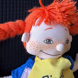 May include: A handmade doll with vibrant orange yarn pigtails and freckles. The doll is dressed in a blue shirt and a yellow apron with the name "Pippi" printed on it. The doll has a smiling expression.