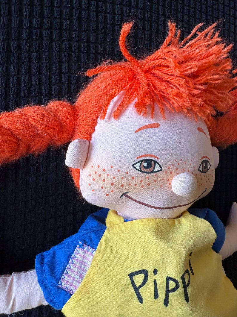 May include: A handmade doll with vibrant orange yarn pigtails and freckles. The doll is dressed in a blue shirt and a yellow apron with the name "Pippi" printed on it. The doll has a smiling expression.
