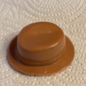 May include: A small, brown plastic hat with a rounded crown and a wide brim. The hat has a ridged band around the base of the crown. The hat is sitting on a white, textured surface.