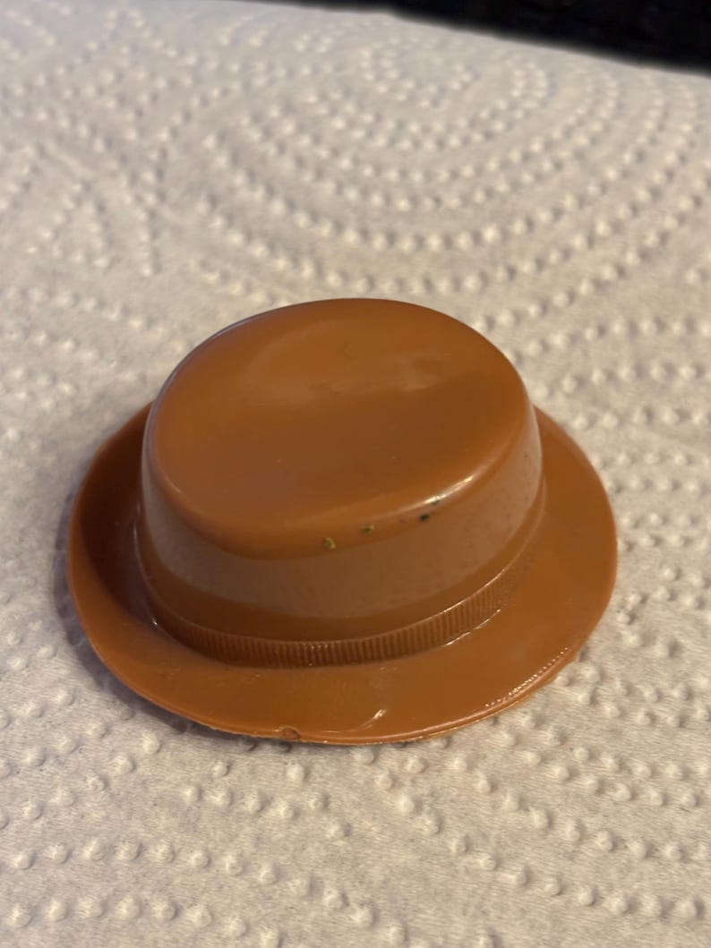 May include: A small, brown plastic hat with a rounded crown and a wide brim. The hat has a ridged band around the base of the crown. The hat is sitting on a white, textured surface.