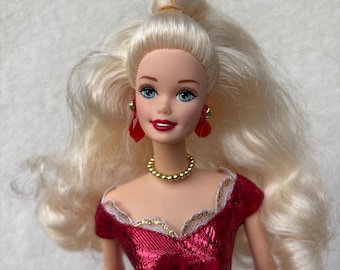 May include: A doll with long, wavy blonde hair, blue eyes, and red lipstick. She wears a red dress with a gold-colored necklace and matching earrings. The dress has a white lace trim and a white and gold-colored purse.