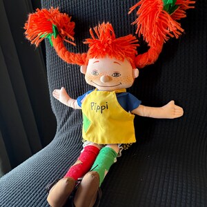 May include: A Pippi Longstocking doll with vibrant orange yarn pigtails secured with green bows. The doll is dressed in a blue top, a yellow apron bearing the name "Pippi", and mismatched red and green socks. Brown shoes complete the ensemble.