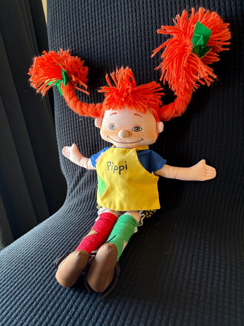 May include: A Pippi Longstocking doll with vibrant orange yarn pigtails secured with green bows. The doll is dressed in a blue top, a yellow apron bearing the name "Pippi", and mismatched red and green socks. Brown shoes complete the ensemble.