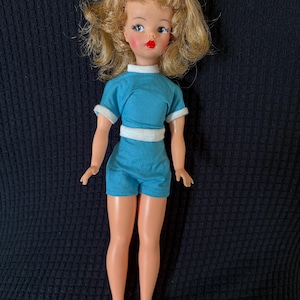 Könnte beinhalten: Eine Vintage-Puppe mit blondem Haar, blauen Augen und roten Lippen. Die Puppe trägt ein türkisblaues Oberteil und Shorts mit weißem Besatz sowie weiße Schuhe. Die Puppe steht vor einem schwarzen Hintergrund.