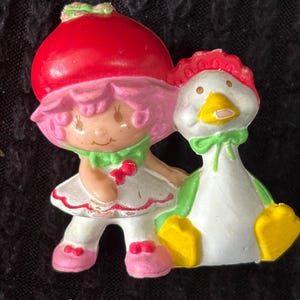 May include: A vintage Strawberry Shortcake figurine with a red strawberry hat and pink hair, holding a white and green duck. The duck has yellow feet and a green bow. The figurine is a collectible item.