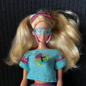 May include: A vintage doll with long blonde hair, wearing a teal crop top and skirt with pink accents. The doll has blue goggles, a pink and blue headband, and a pink watch. The top features a circular graphic.