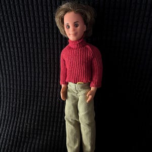 May include: A vintage doll wearing a red turtleneck sweater, light green pants, and brown shoes. The doll has brown hair and is standing against a black textured background. The doll's outfit includes a gold-colored belt buckle.