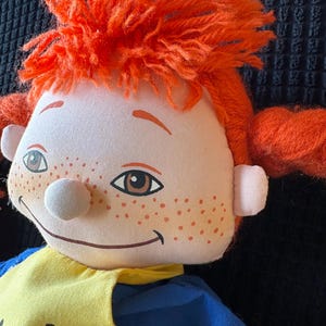May include: A soft doll with a smiling face, freckles, and orange yarn hair in pigtails. The doll is dressed in a yellow and blue outfit. The doll's face features brown eyes and a white nose.