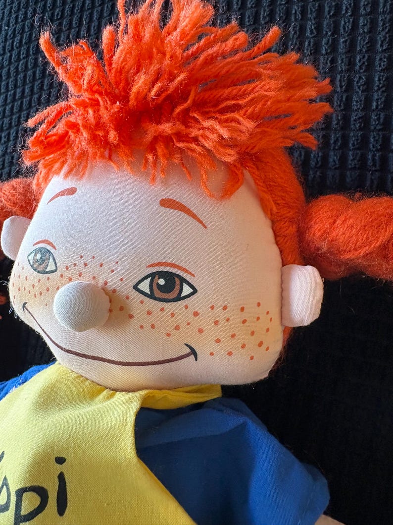 May include: A soft doll with a smiling face, freckles, and orange yarn hair in pigtails. The doll is dressed in a yellow and blue outfit. The doll's face features brown eyes and a white nose.